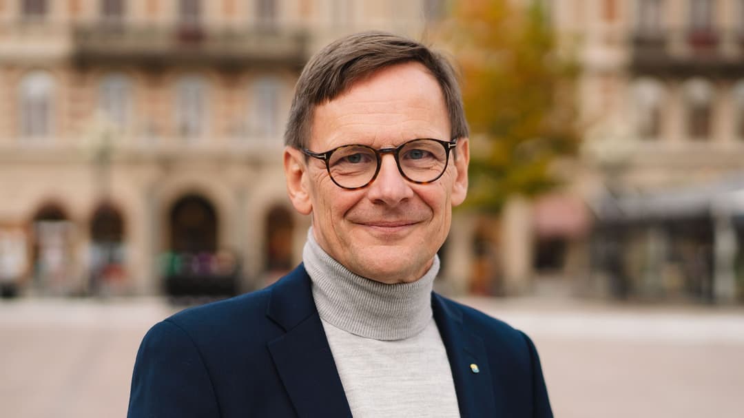 En mörkhårig man med glasögon, grå polotröja och mörk kavaj står på ett torg och tittar in i kameran. 