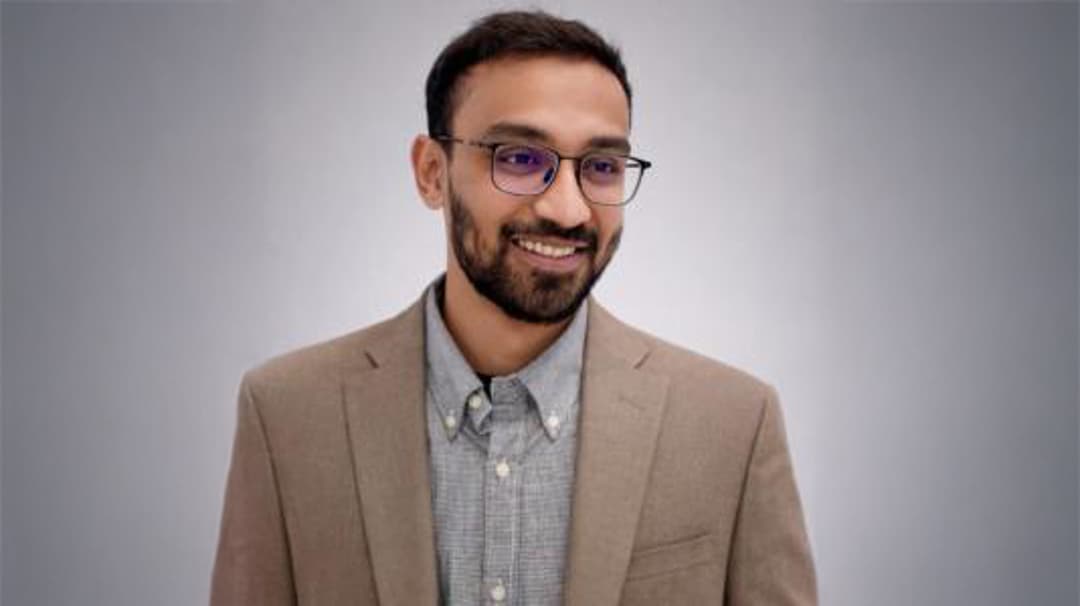 A smiling man with a beige jacket and glasses