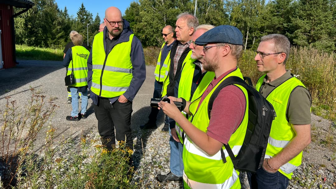Several people in neon yellow vests stand together in a group outdoors. They look towards a red building in the background. One person holds a measuring tool in his hand.