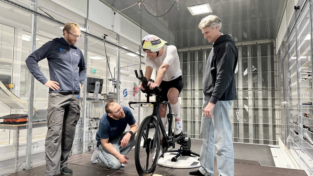 Tre män står och tittar på en fjärde man som sitter på en cykel inomhus.