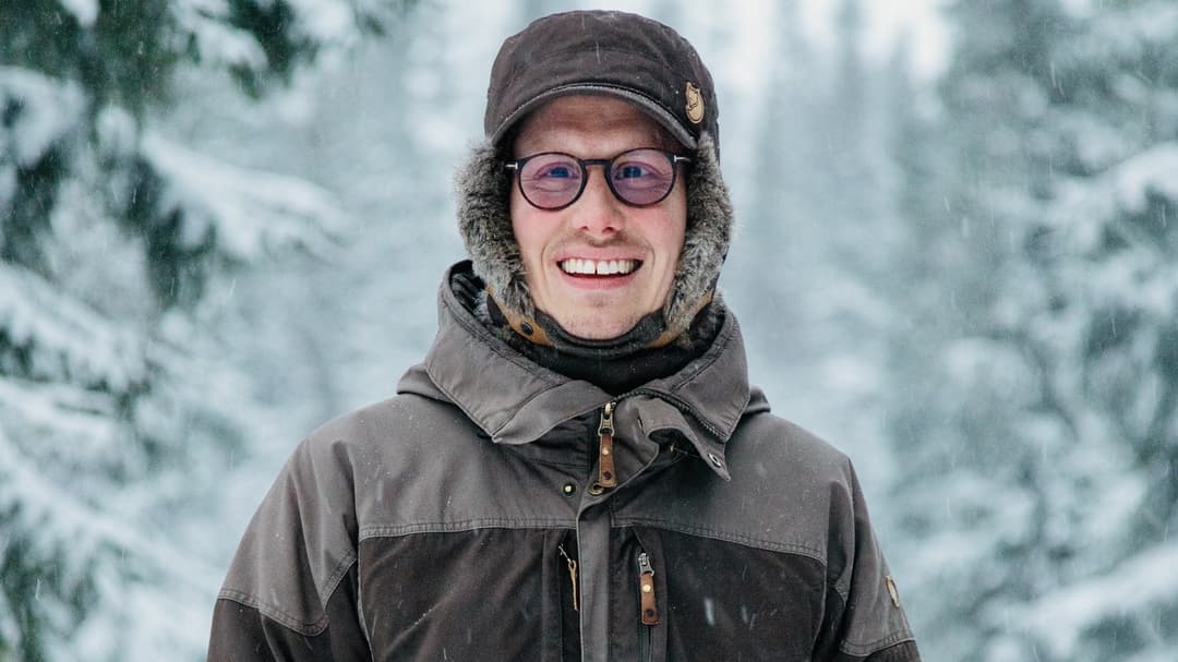 Glad ung man i tjocka vinterkläder står utomhus i vinterlandskap med snöklädda granar i bakgrunden