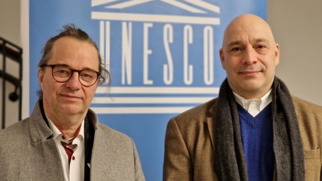 Two men dressed in coats stand in front of a blue sign with the UNESCO logo 