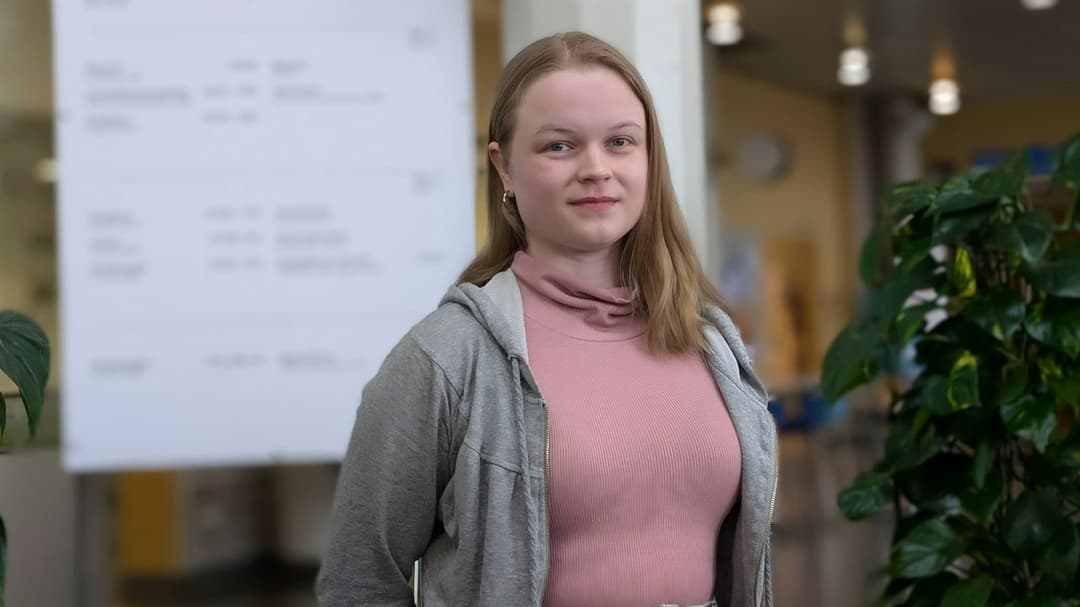 En kvinna med långt hår står framför en ljustavla. Bredvid finns en grön växt.