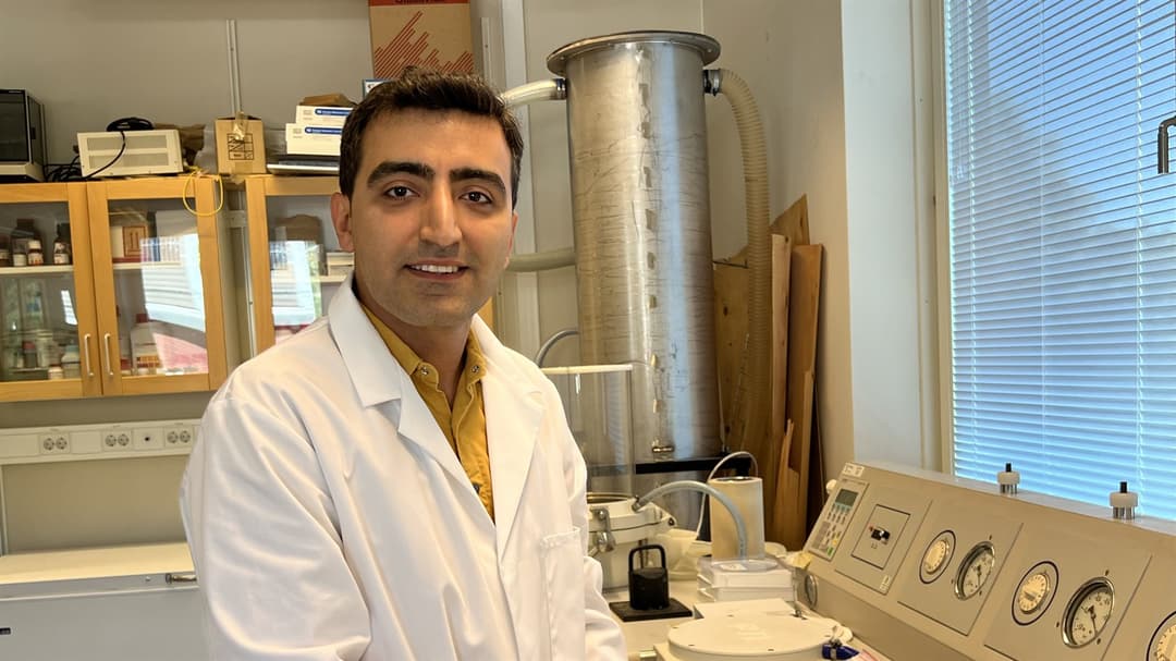  A man in a white coat in a research laboratory looks into the camera and smiles.