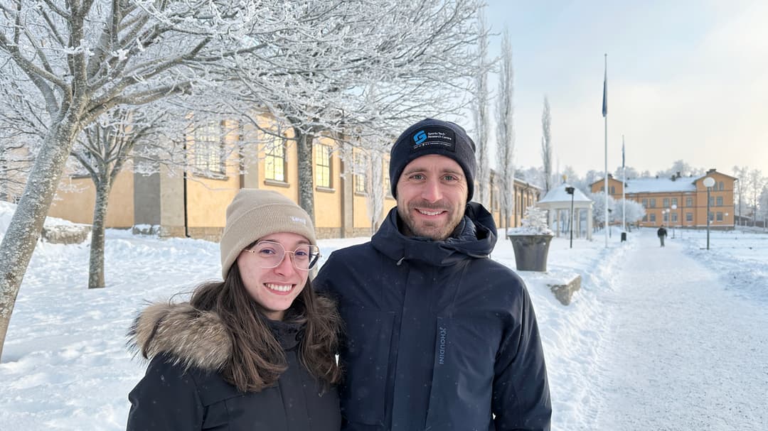 Två personer står framför frostiga träd och byggnader klädda i vinterkläder.