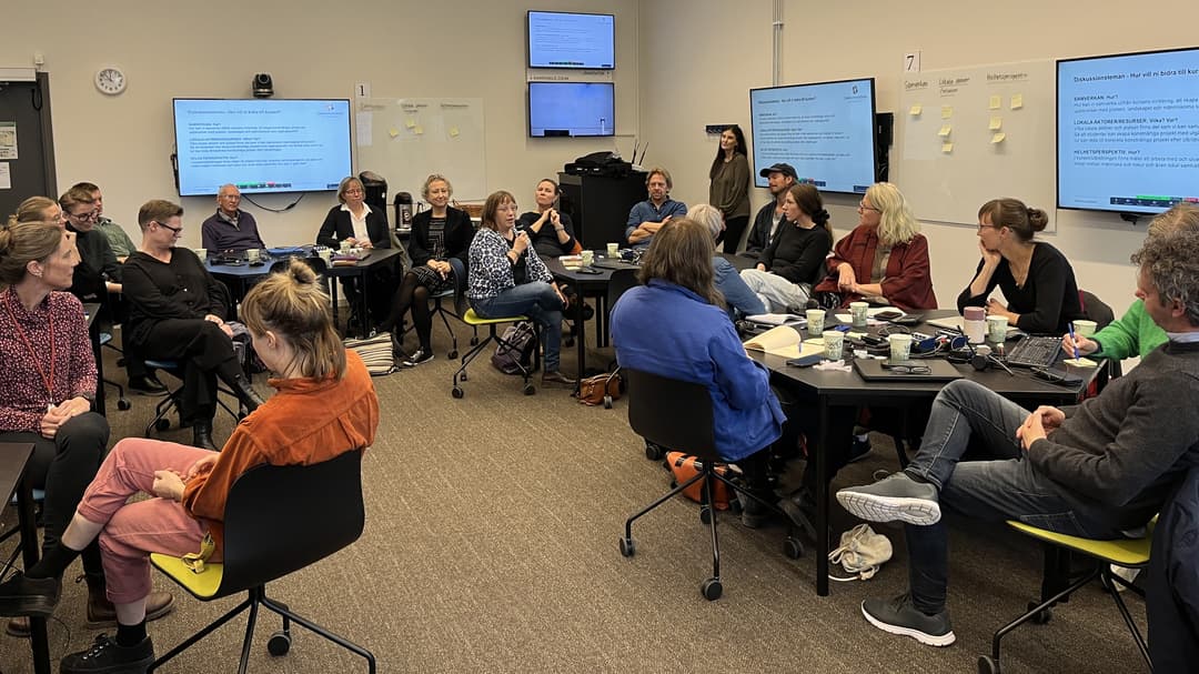 About thirty people sit at round tables in a room and talk in small groups. 