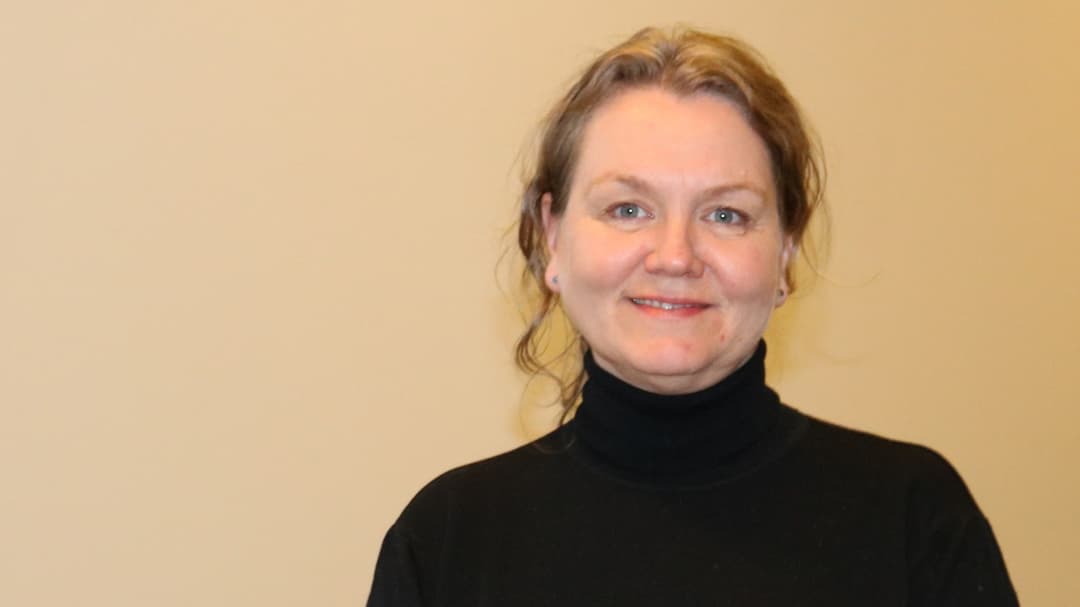 A woman with brown hair tied up and wearing a black polo shirt in a corridor with beige walls.