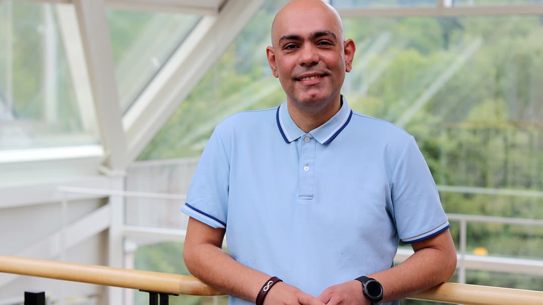 Man in blue short-sleeved shirt stands by a railing and smiles at the camera.