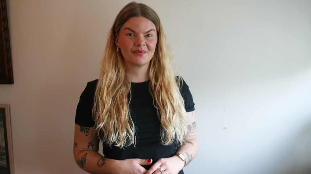 Kvinna i svart kortärmad tröja och långt blont hår står framför en beige vägg. 
