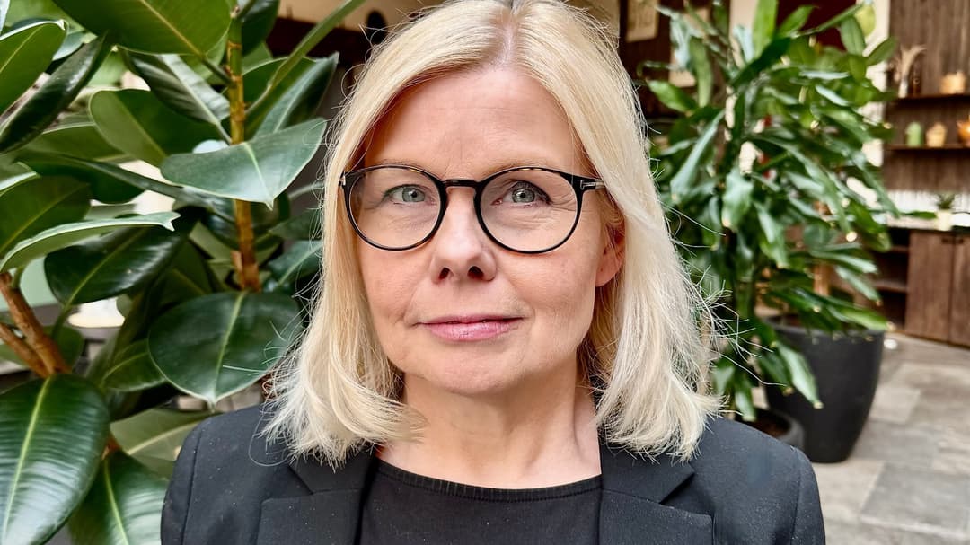 En kvinna med axellångt blont hår och glasögon, klädd i svart kavaj står framför gröna, höga växter i inomhusmiljö. 