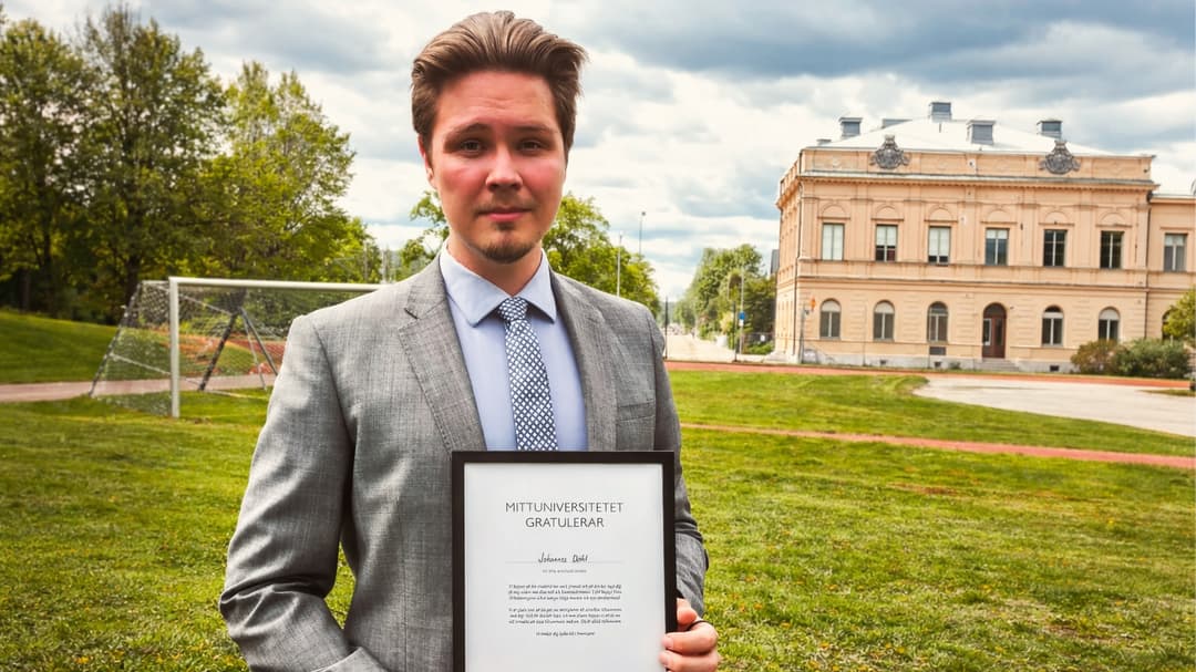 En man i grå kostym håller i ett diplom. I bakgrunden finns en gräsmatta och en byggnad.