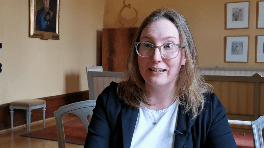 A woman with long brown hair down to her shoulders and wearing a black jacket is interviewed.