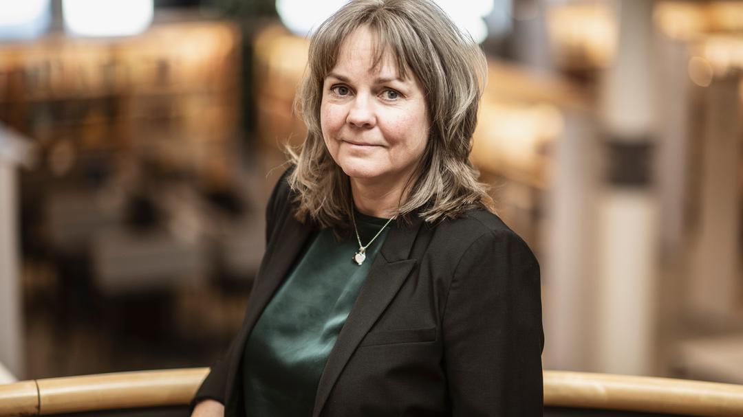 En kvinna i ljusbrunt hår, grön blus och svart kavaj står i biblioteksmiljö.