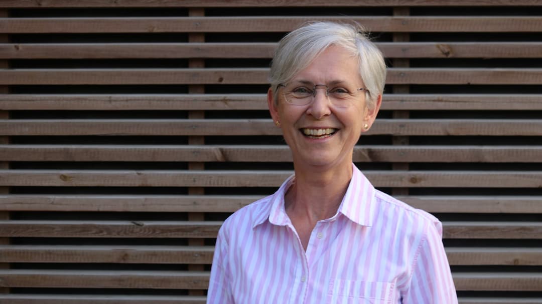 A woman with grey short hair, glasses and a pink and white striped shirt stands in front of a wall with horizontal wooden panelling.