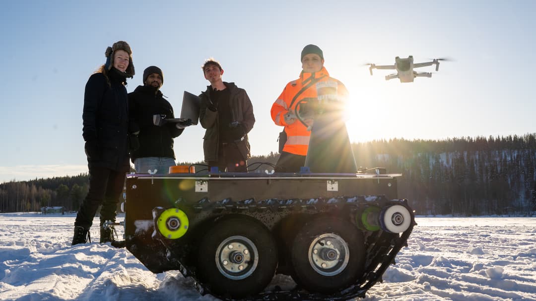  Four people are standing in a snowy landscape. A drone flies behind them and a small all-terrain vehicle stands in front of them.