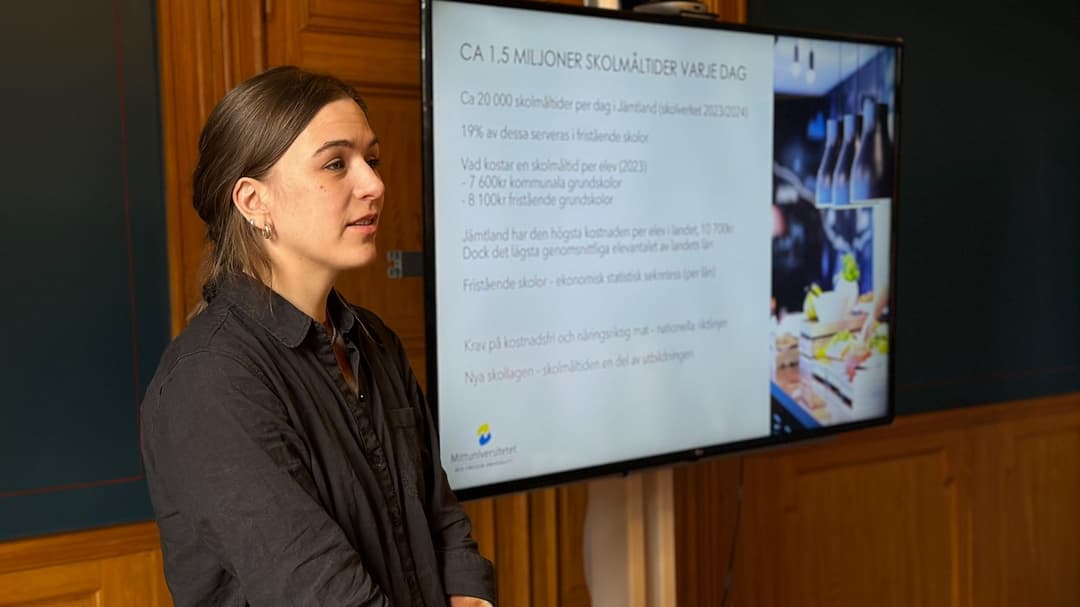 A woman with brown hair and a dark grey shirt stands by a screen with a presentation on it.