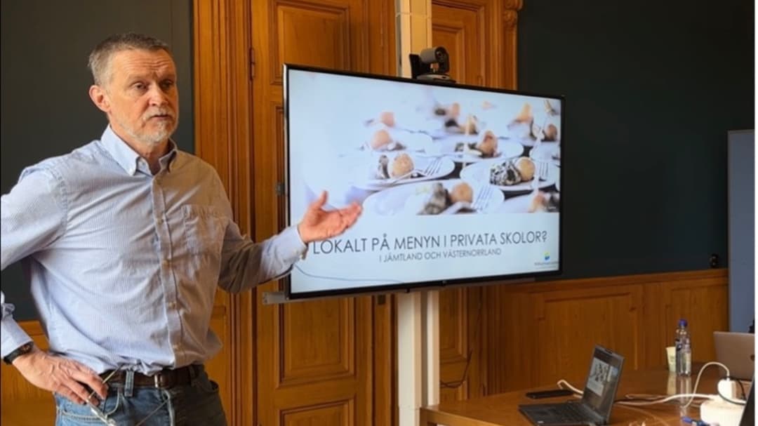 A man in a light blue shirt stands in front of a screen with a presentation on it and gestures with his left hand.