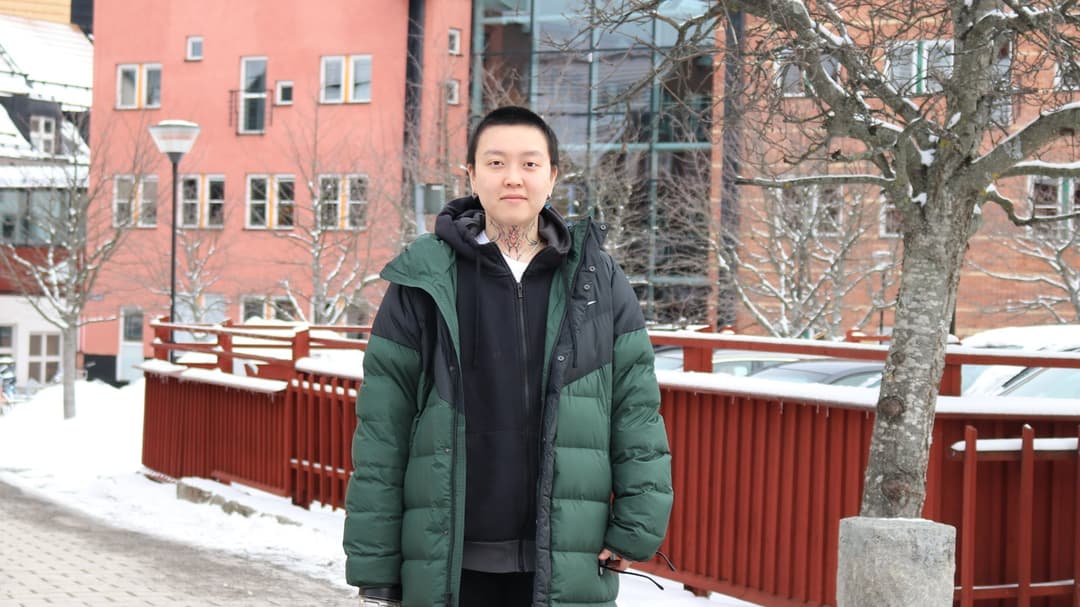 En person i en grön jacka står framför ett träd, med flera byggnader i bakgrunden.