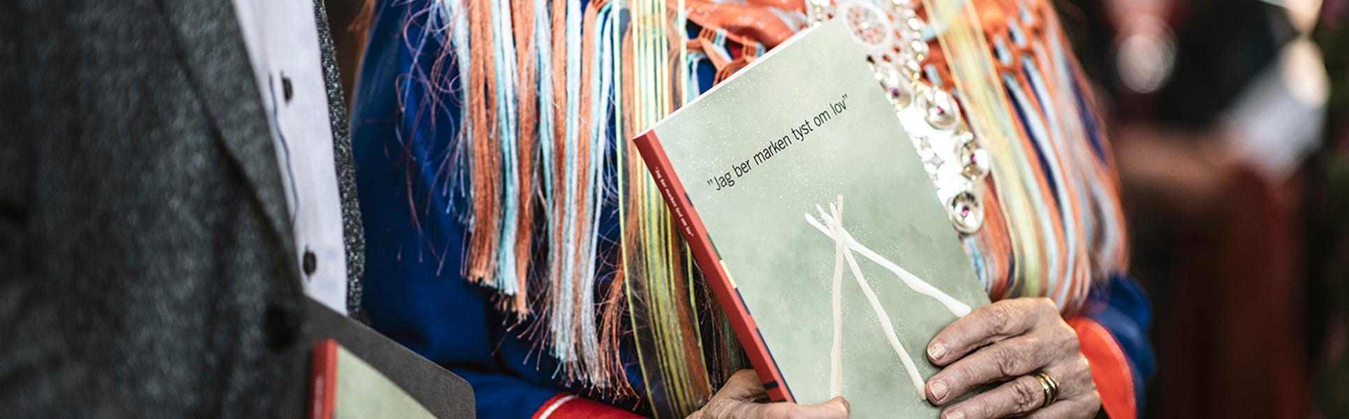 En kvinna i samisk folkdräkt håller i en bok som har rubriken "Jag ber marken tyst om lov".
