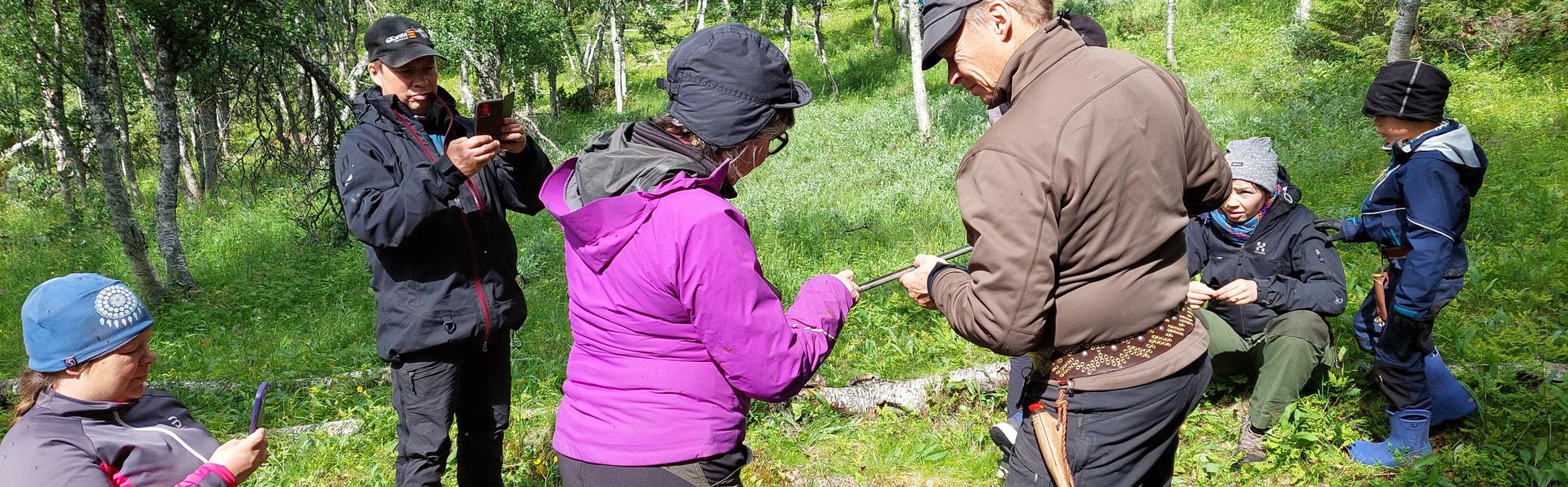 Två personer studerar ett jordprov i en jordsond i en fjällbjörkskog. Fyra personer ser på. 