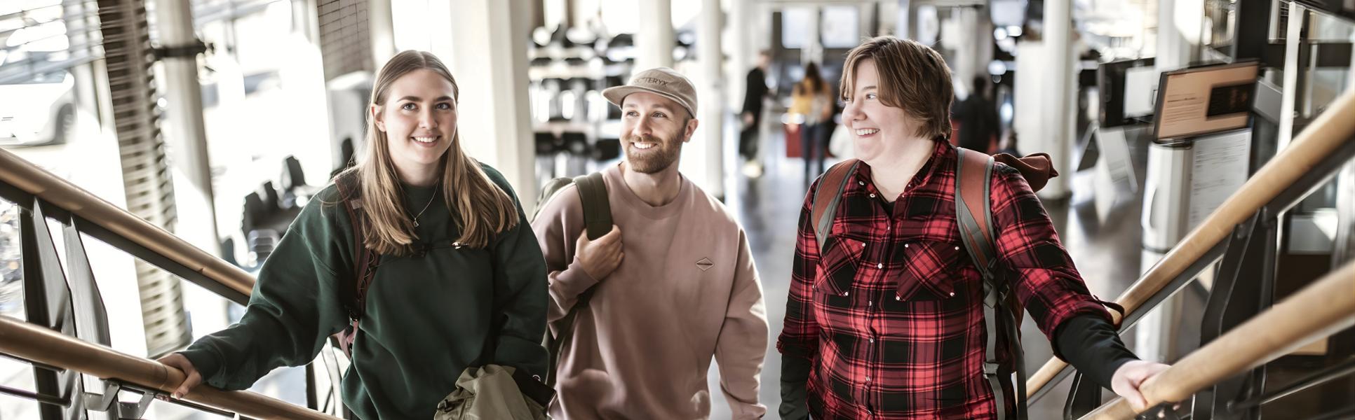 Tre unga studenter står i en trappa. De är alla glada. 