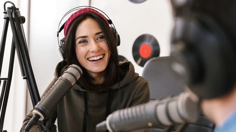 En tjej och en kille pratar med varandra i en radiostudio. Tjejen är vänd mot kamera och skrattar. Killen ser man bakifrån. De pratar i mikrofoner. Väggen i bakgrunden är dekorerad med LP-skivor.
