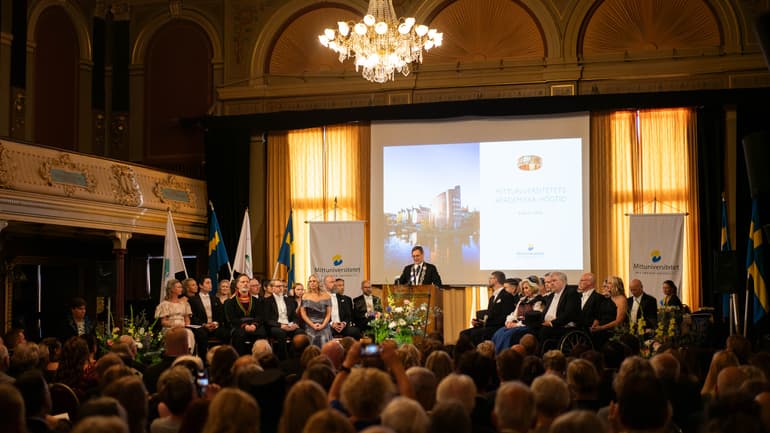En finklädd man står i en talarstol och talar framför en publik.