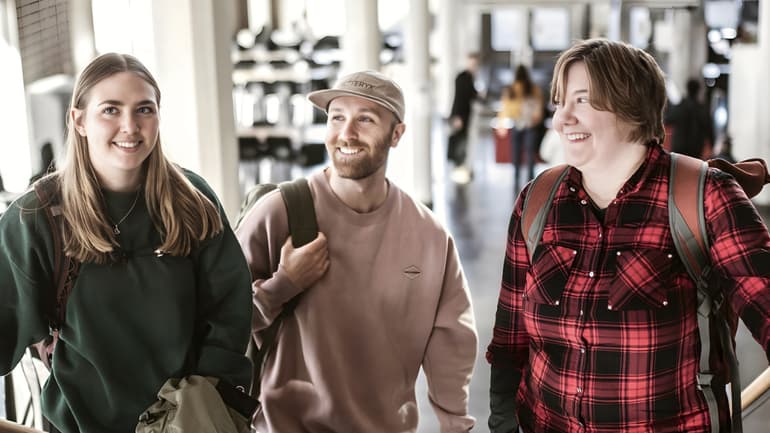 Tre unga studenter står i en trappa. De är alla glada. 