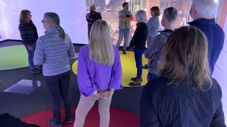 En grupp människor står i ett rum med en stor skärm som visar en stadsmiljö.