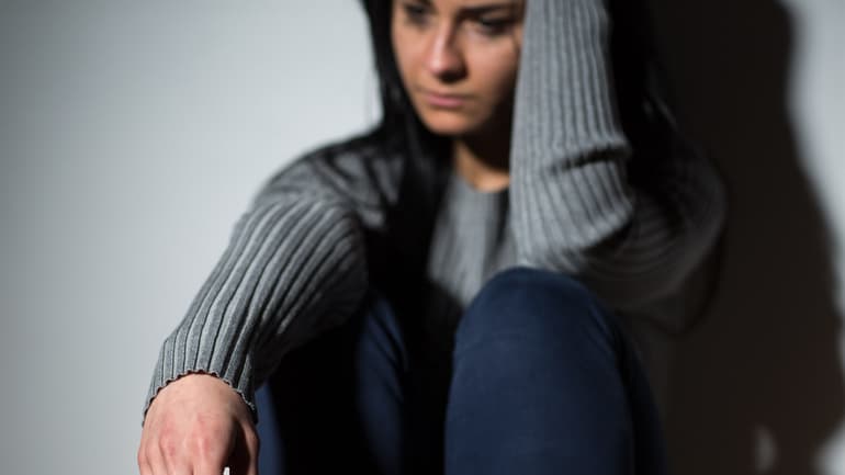 Woman wearing jeans and a gray sweater sits leaning against a wall and looks sad.