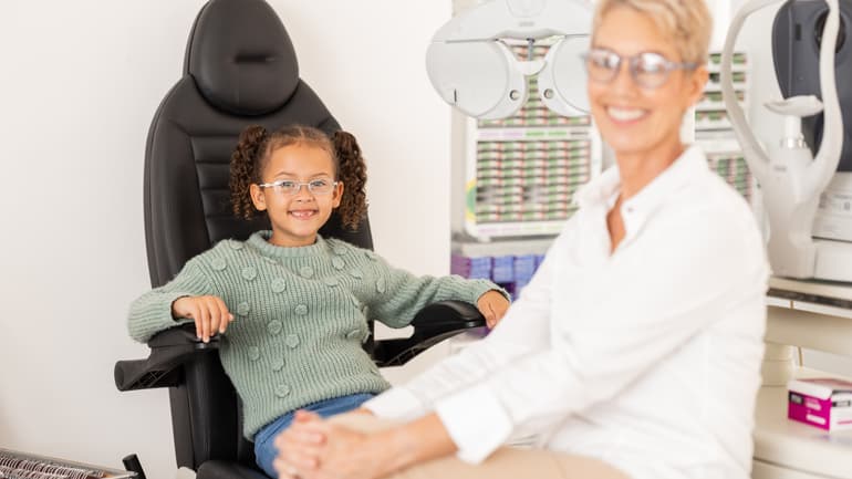 A child wearing glasses is sitting in an optician's chair. A woman is sitting in front of her. Both are smiling.