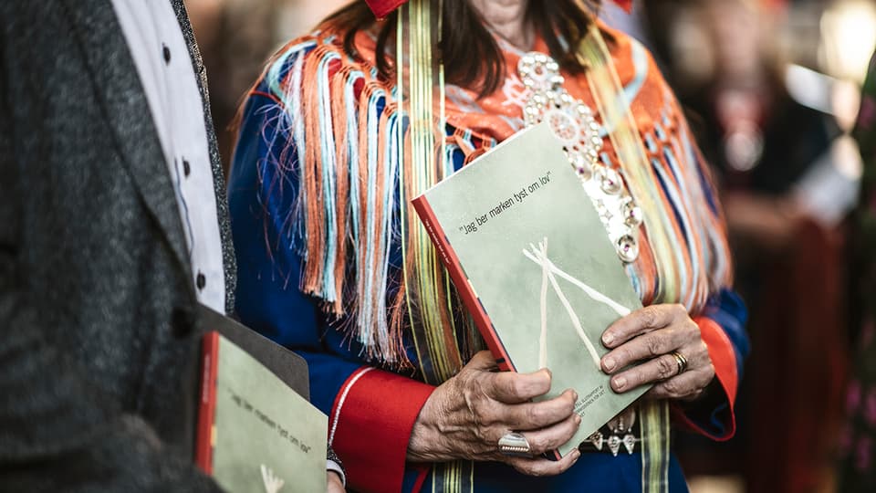 A woman wearing traditional Sámi dress is holding a book titled I Silently Ask the Land for Permission.