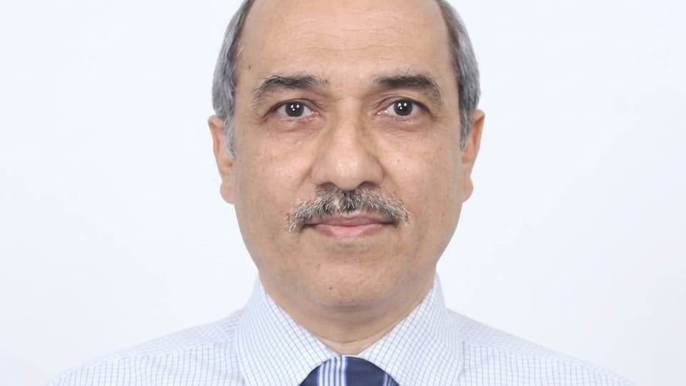 A close-up of a brown-eyed man with greying hair and a moustache. He is wearing a shirt and tie and looks serious.