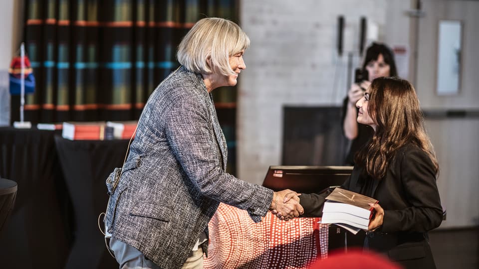 Kvinna med ljust hår, klädd i grå kavaj, skakar hand med en kortare kvinna med mörkt hår, mörk kavaj, som håller i sanningskommissionens slutbetänkande.