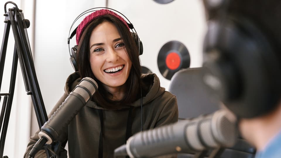 En tjej och en kille pratar med varandra i en radiostudio. Tjejen är vänd mot kamera och skrattar. Killen ser man bakifrån. De pratar i mikrofoner. Väggen i bakgrunden är dekorerad med LP-skivor.