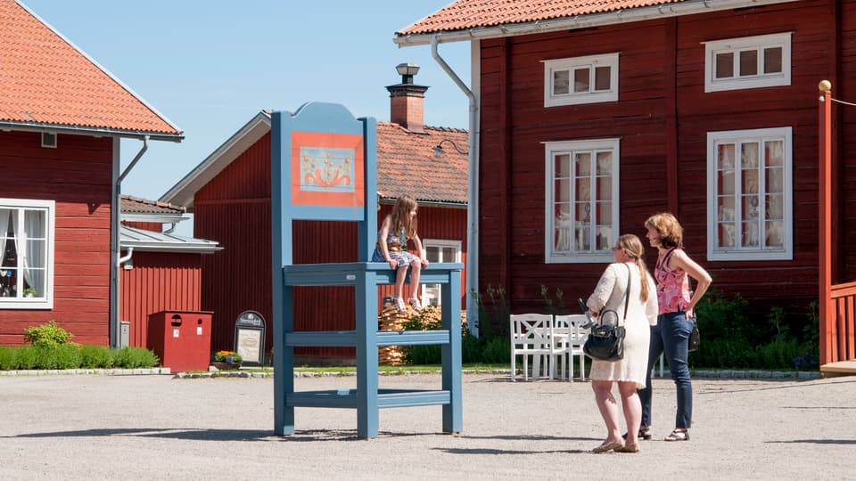 A girl sits on a large chair outside three red houses and two women stand below and look at the girl.