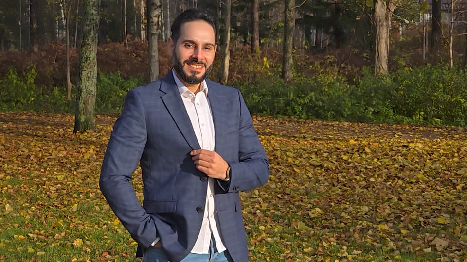 A man looks into the camera and smiles, in the background there are trees and yellow leaves on the ground.