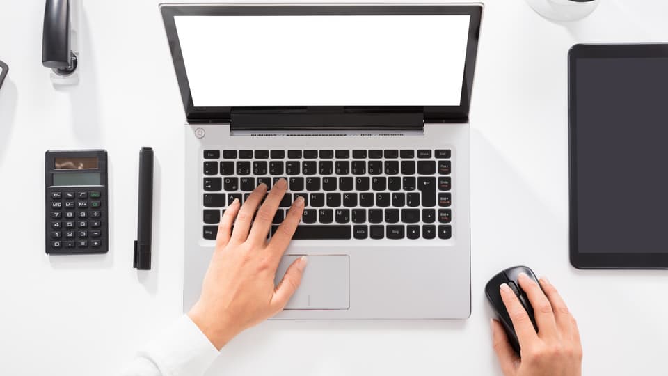 Händer knappar på en laptop, miniräknare, tuschpenna och häftmaskin ligger in till