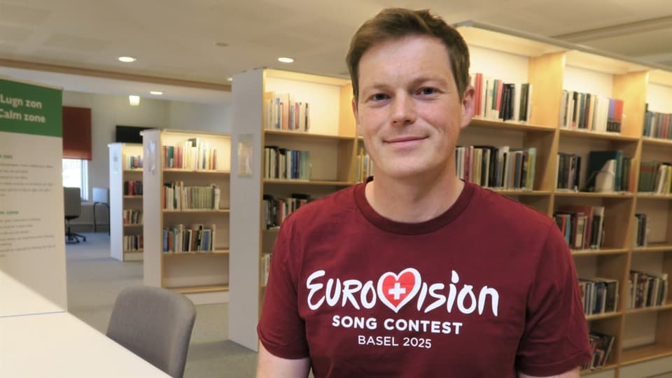 Young man with a short haircut in a dark red t-shirt with Eurovision print, sitting at a table in a library with books in the background