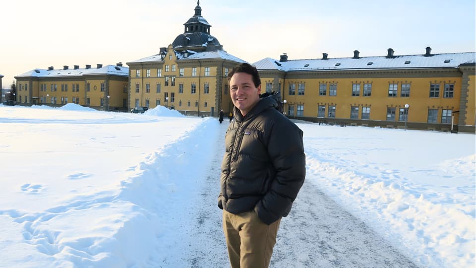 Man i svart dunjacka står framför byggnaden klockhuset på Mittuniversitetets campus i Östersund. Han ser glad ut. 
