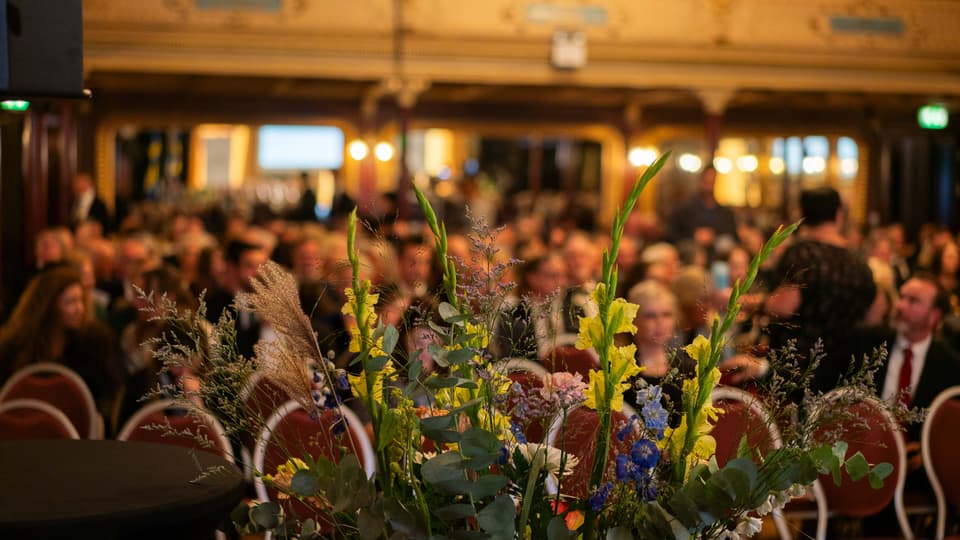 Blomsterarrangemang på en scen med uppradade stolar i bakgrunden.
