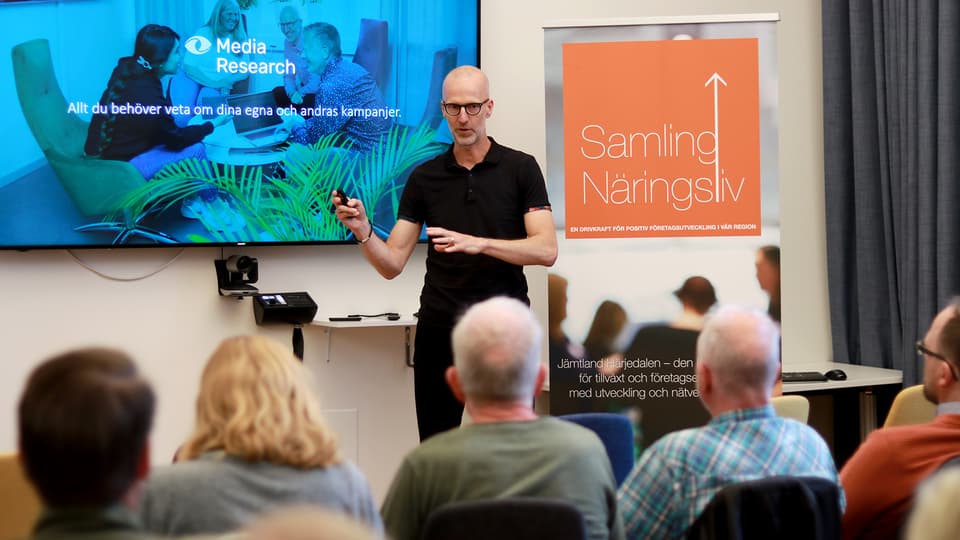 En man som står bredvid en stor skärm och talar för en sittande publik