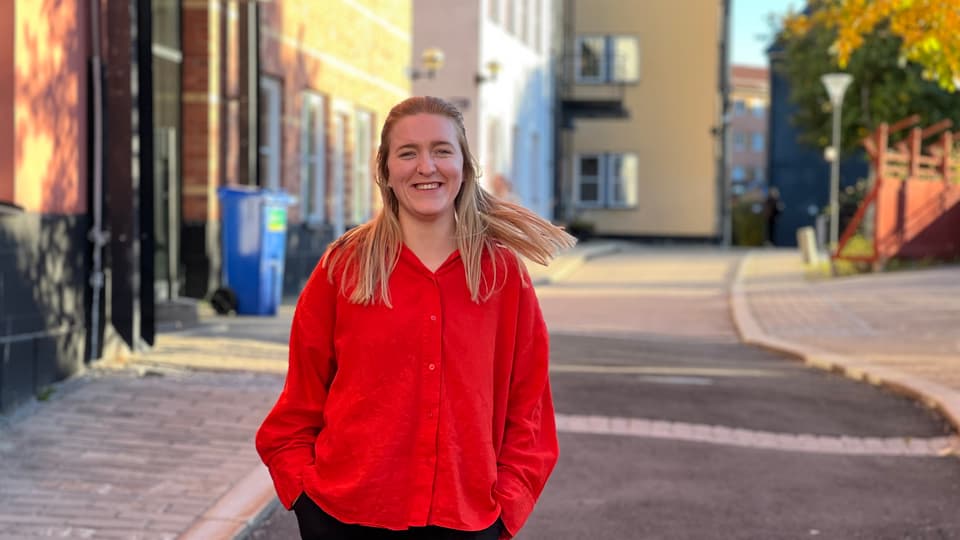 A woman in a red blouse looks into the camera and smiles. She has a street and some house facades in the background.