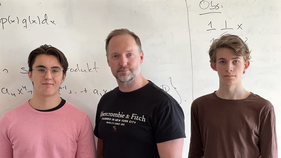 Two younger men and a middle-aged man stand in front of a whiteboard.