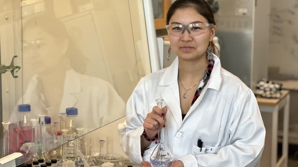 A woman in a white coat and protective glasses stands in a laboratory.