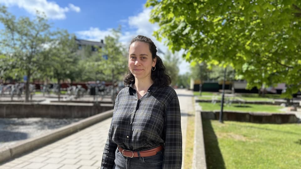 A woman is standing on a cobblestone path, looking directly at the camera. Trees can be seen in the background.