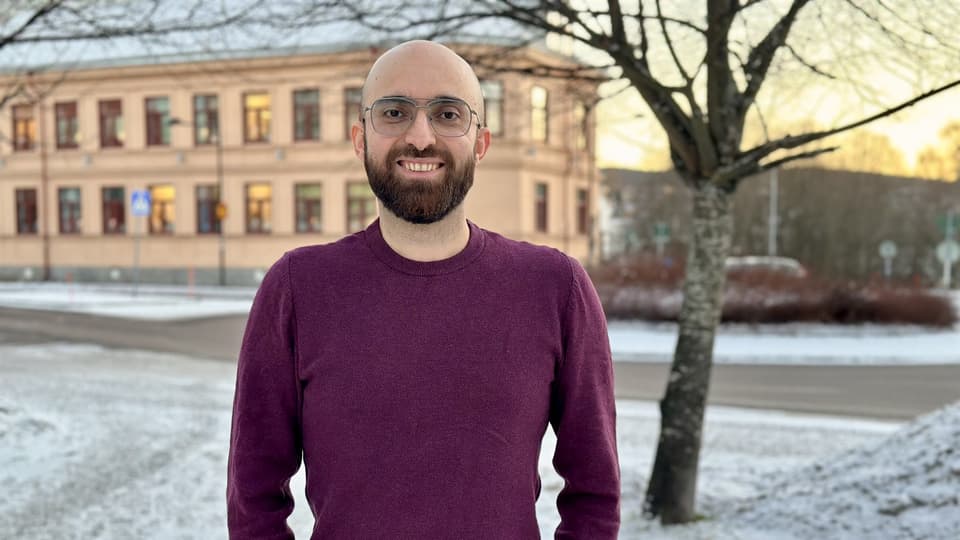 Smiling man with a shaved head, brown beard, and glasses. He is wearing a burgundy sweater and standing outdoors. The season is winter.