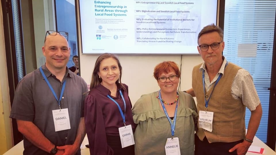 Four people, two men and two women, are standing in a room in front of a screen with a presentation on it. Around their necks are blue lanyards with name tags.