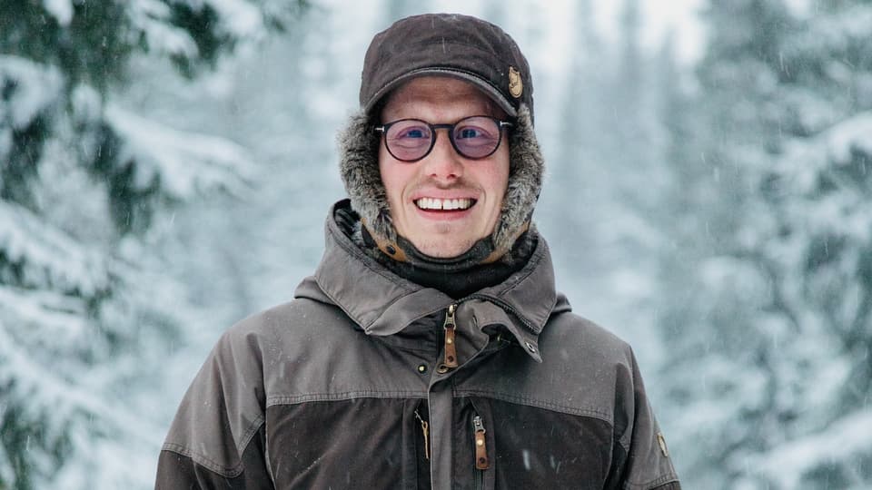 Happy young man in thick winter clothes standing outdoors in a winter landscape with snow-covered fir trees in the background.