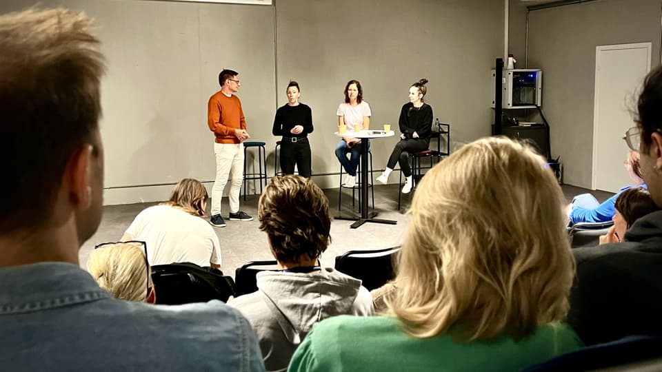 Four people are standing at a round table and talking to an audience.